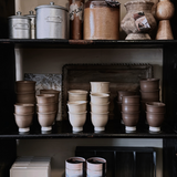 A shelf filled with various handmade pottery items, including cups and vases, with a soft matte finish in beige, brown, and gray colors.