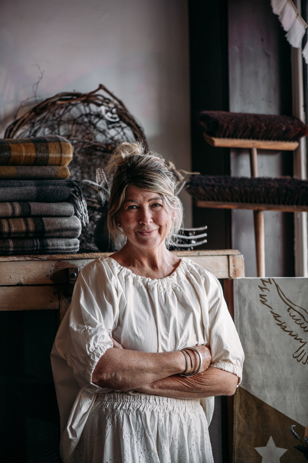 Belinda Satterthwaite (The Shopkeeper) standing in a rustic interior setting with wooden furniture and decorative items.
