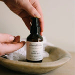 Hands dispensing Olieve & Olie Lemon Scented Gum Hand Sanitizer from an amber pump bottle resting in a shallow ceramic bowl.