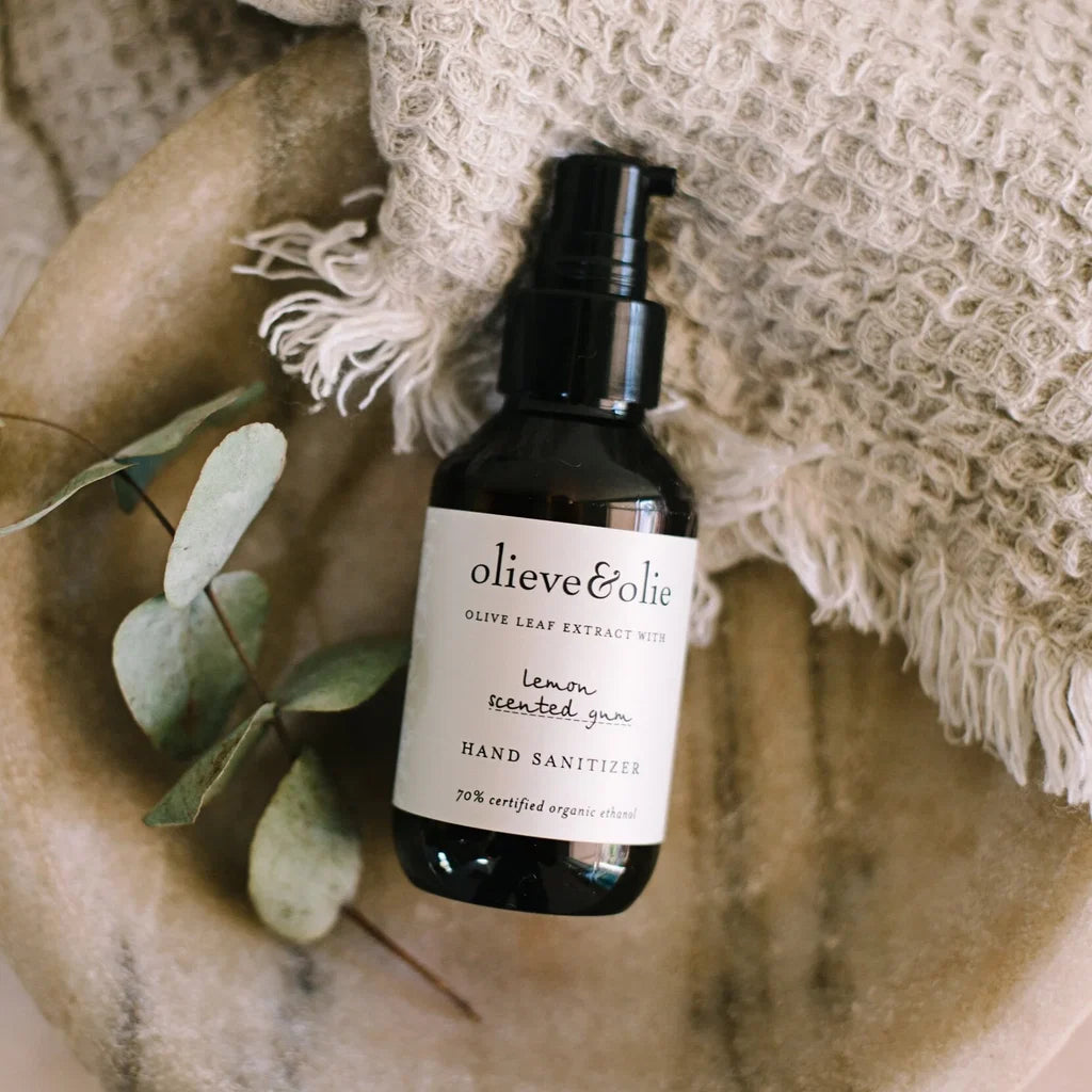 Olieve & Olie Lemon Scented Gum Hand Sanitizer in an amber pump bottle, styled with eucalyptus leaves and a textured natural-fibre cloth.
