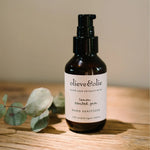 Amber bottle of Olieve & Olie Lemon Scented Gum Hand Sanitizer with 70% certified organic ethanol, styled beside eucalyptus leaves on a wooden surface.