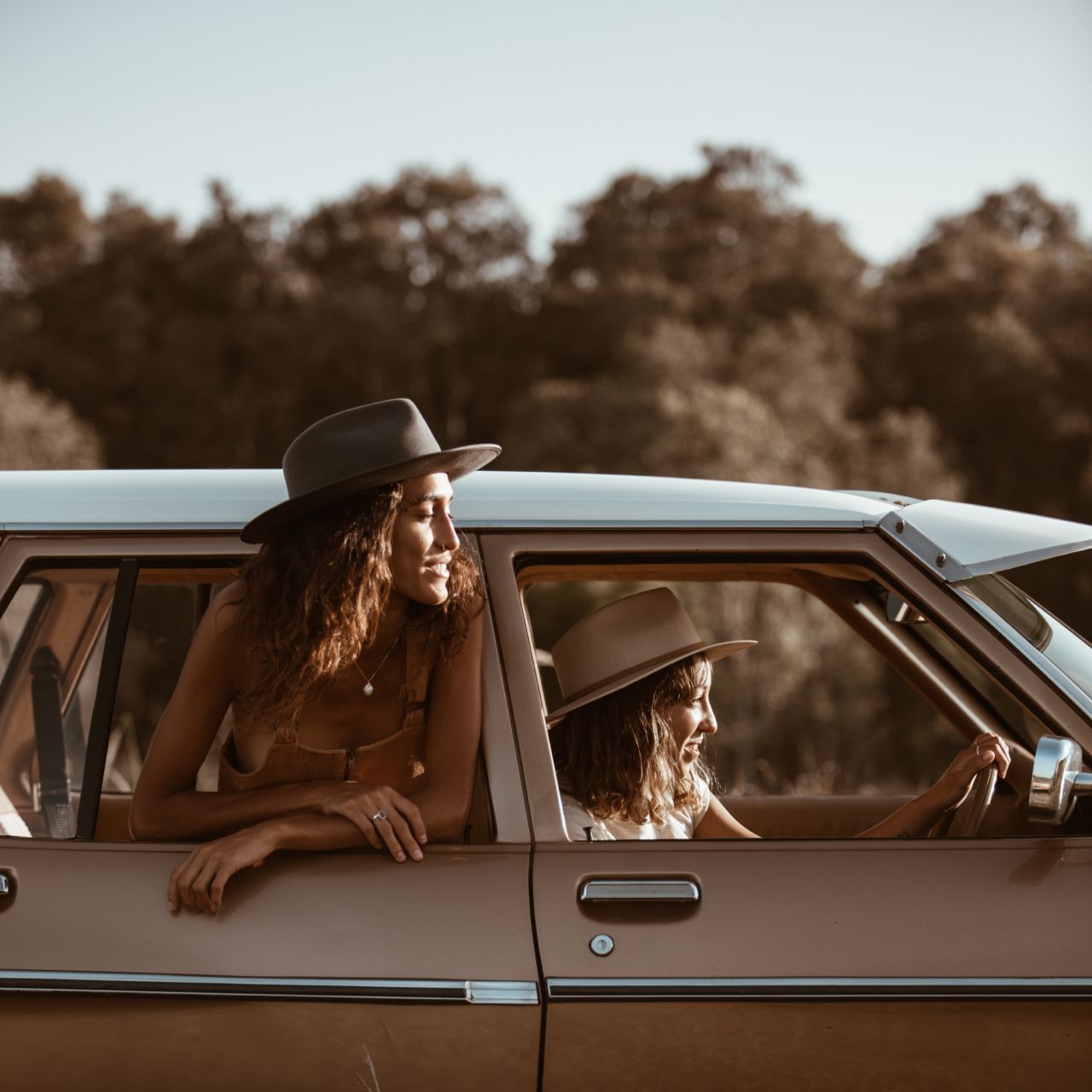 Two women smiling at the sunset from their car wearing Will & Bear hats